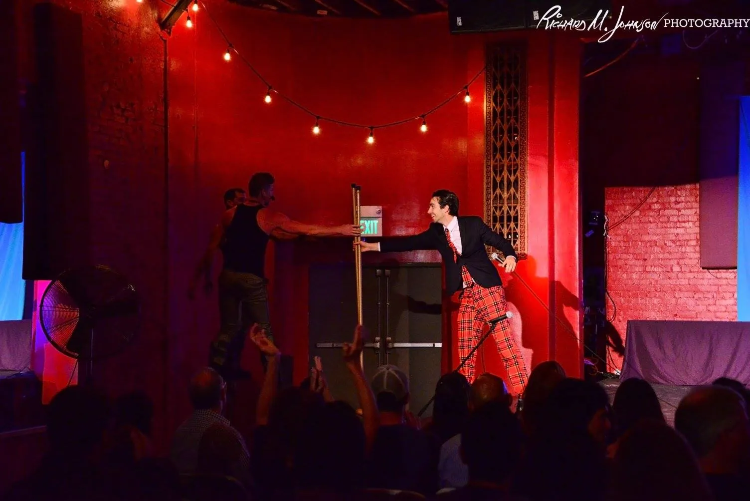 Performers on stage under string lights and red brick, with a packed audience